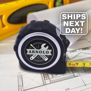 May include: A black tape measure with a circular center emblem featuring a screwdriver and wrench graphic with the name ARNOLD printed across a banner. The tape measure extends to show a yellow blade with black markings and the text 5m.