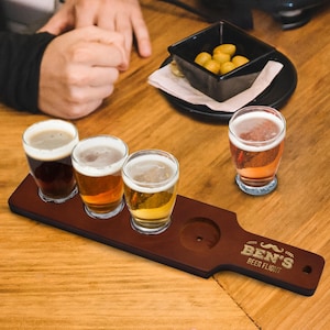 May include: A wooden beer flight paddle with four glasses of beer. The paddle is engraved with "BEN'S BEER FLIGHT" and a mustache design. The beers vary in color from dark brown to light gold. A small black bowl of snacks sits nearby.