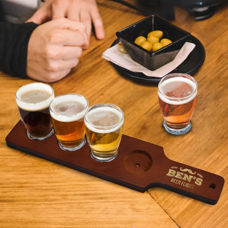 May include: A wooden beer flight paddle with four glasses of beer. The paddle is engraved with "BEN'S BEER FLIGHT" and a mustache design. The beers vary in color from dark brown to light gold. A small black bowl of snacks sits nearby.