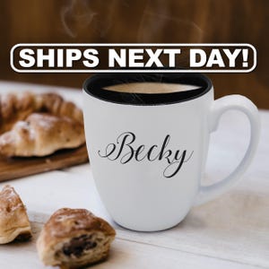 Pu&ograve; includere: Una tazza in ceramica bianca con interno e manico neri, caratterizzata dal nome Becky stampato in un carattere corsivo nero sulla parte anteriore. La tazza &egrave; riempita con un liquido scuro e mostra volute di vapore che salgono dal bordo.
