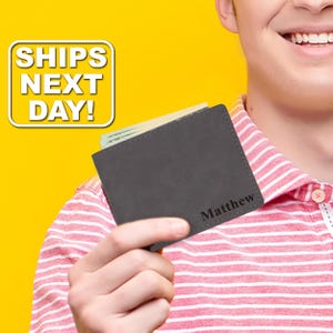 May include: A dark gray bifold wallet featuring the name Matthew engraved in a black serif font on the bottom right corner. The wallet is constructed with a smooth matte finish and contains several paper bills tucked inside the main compartment.