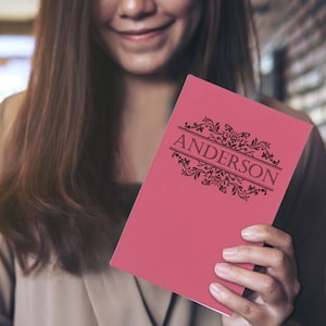 May include: A pink notebook with the name "ANDERSON" in black, stylized text. The text is framed by decorative floral elements. The notebook is held by a person.