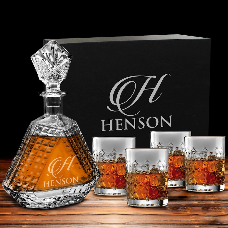 May include: A clear glass decanter with a diamond-cut pattern and a matching stopper, accompanied by three matching whiskey glasses. The decanter and the black presentation box behind it feature the letter H and the text HENSON.
