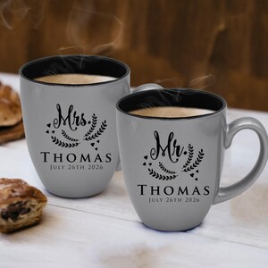 May include: Two grey ceramic mugs with black interiors, filled with a warm beverage. One mug is labelled "Mrs. THOMAS JULY 26TH 2026" and the other "Mr. THOMAS JULY 26TH 2026", with decorative floral elements. Steam rises from the mugs.