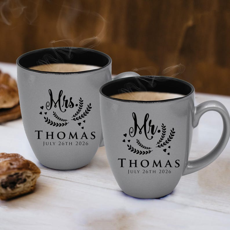 May include: Two grey ceramic mugs with black interiors, filled with a warm beverage. One mug is labelled "Mrs. THOMAS JULY 26TH 2026" and the other "Mr. THOMAS JULY 26TH 2026", with decorative floral elements. Steam rises from the mugs.