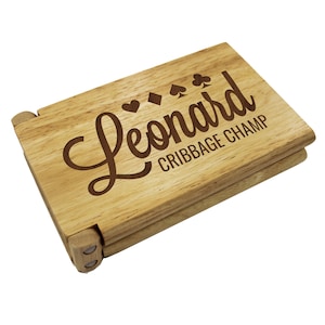 Puede incluir: Un tablero de cribbage de madera con el nombre "Leonard" y el texto "Cribbige Champ" grabado en &eacute;l. El tablero est&aacute; cerrado y tiene un color marr&oacute;n claro.