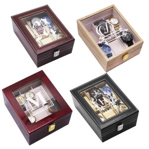 May include: Four watch display cases in various colors: dark brown, light brown, dark red, and black. Each case has a glass top, a beige interior, and a silver clasp. The cases are personalized with names or initials.