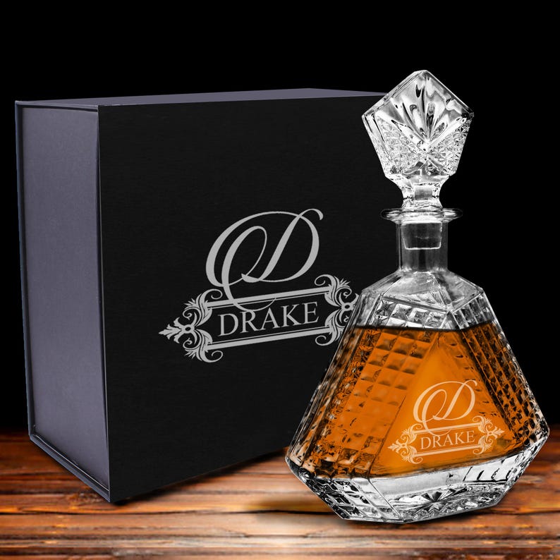 May include: A clear, faceted glass decanter filled with amber liquid, featuring an etched letter D and the name DRAKE. The decanter has a geometric stopper and sits beside a black gift box displaying the same etched logo and name DRAKE.