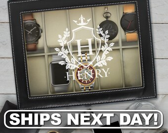 May include: A black rectangular watch display case with a glass lid featuring a white printed crest with the letter H and the name HENRY. The interior contains beige velvet cushions holding various wristwatches and a pocket watch.