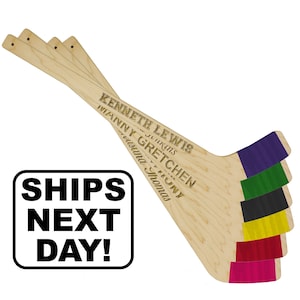 May include: A set of five light-colored wooden hurling sticks, each featuring a different colored tape at the base in purple, green, black, yellow, red, and pink. The sticks are engraved with various names including Kenneth Lewis and Gretchen.