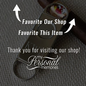 May include: Silver cigar cutter with a personalized inscription. The cutter has a round blade and a lever to open and close the blade. The cutter is on a brown background.