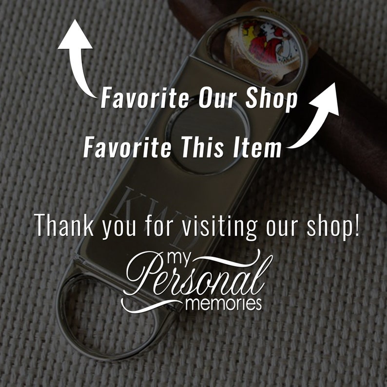 May include: Silver cigar cutter with a personalized inscription. The cutter has a round blade and a lever to open and close the blade. The cutter is on a brown background.