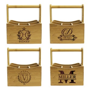 Może przedstawiać: Cztery drewniane przenośne pojemniki na butelki z uchwytami. Każdy pojemnik ma inny grawerowany wz&oacute;r z nazwą i inicjałem. Wzory obejmują herb z literą H dla Henry'ego, monogram z literą D dla Drake'a, herb z literą V dla Victorii i duże M dla Millera.