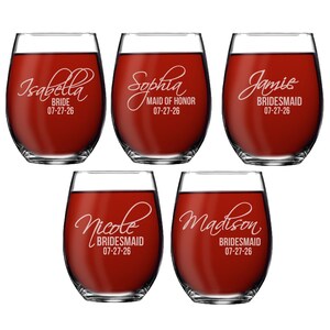 May include: Five stemless wine glasses filled with red liquid. Each glass has a name and role, such as "Isabella Bride" and "Sophia Maid of Honour", with the date "07-27-26" below. The glasses are clear with white lettering.