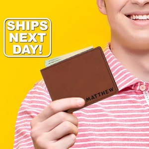 May include: A brown leather bifold wallet featuring the name MATTHEW engraved in black lettering on the bottom right corner. The wallet is shown holding paper currency inside its main compartment, with visible dark stitching along the edges.