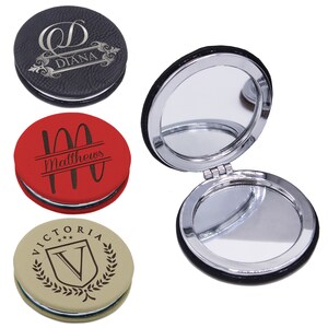 May include: Three round compact mirrors with textured covers in black, red, and tan, each featuring custom laser-engraved text and designs. One mirror is shown open, revealing two reflective surfaces inside a polished silver-toned metal frame.
