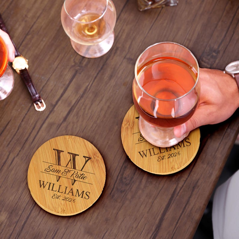 May include: Two round bamboo coasters featuring laser-engraved text. One coaster displays a large letter W, the names Sam & Katie, the surname WILLIAMS, and Est. 2026. The other coaster shows identical text beneath a glass filled with amber liquid.