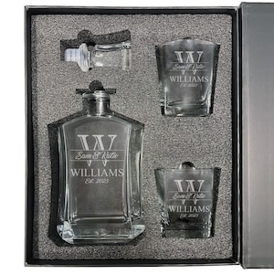 May include: A clear glass decanter and two matching glasses set in a dark gray foam-lined box. Each piece is etched with a large letter W, the names Sam & Katie, the surname WILLIAMS, and the text Est. 2023. A separate glass stopper is included.