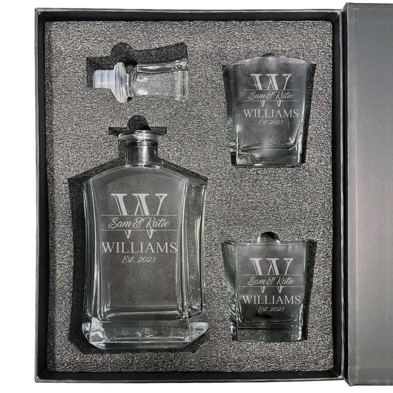 May include: A clear glass decanter and two matching glasses set in a dark gray foam-lined box. Each piece is etched with a large letter W, the names Sam & Katie, the surname WILLIAMS, and the text Est. 2023. A separate glass stopper is included.