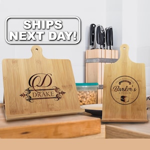 May include: Two light-colored bamboo wood recipe stands with laser-engraved designs. The left stand features a large letter D and the name DRAKE. The right stand displays a circular logo with the text FRESH & DELICIOUS BARKER'S KITCHEN.