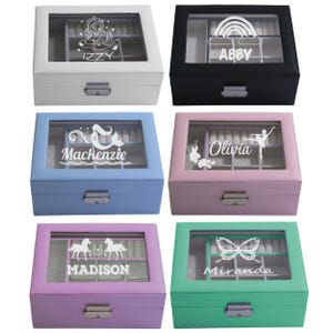 May include: Six rectangular jewellery boxes with glass lids and metal latches in white, black, light blue, pink, purple, and teal. Each lid features a white graphic and a name: Izzy with a unicorn, Abby with a rainbow, Mackenzie with a mermaid, Olivia with a ballerina, Madison with horses, and Miranda with a butterfly.