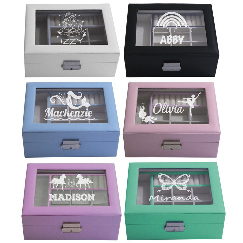 May include: Six rectangular jewellery boxes with glass lids and metal latches in white, black, light blue, pink, purple, and teal. Each lid features a white graphic and a name: Izzy with a unicorn, Abby with a rainbow, Mackenzie with a mermaid, Olivia with a ballerina, Madison with horses, and Miranda with a butterfly.