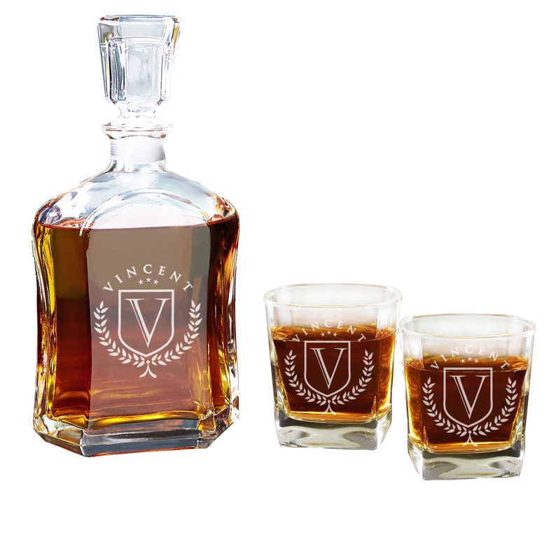 May include: A clear glass decanter with a matching stopper and two square whiskey glasses, all filled with amber liquid. Each piece features an etched design of a shield with the letter V, the name VINCENT, and three stars above it.