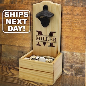 May include: A light-colored wooden wall-mounted bottle opener featuring a black metal opener at the top and a small storage box at the base. The wood is engraved with a large letter M and the name MILLER. The box contains several metal bottle caps.