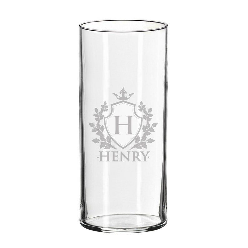 May include: A clear, tall glass with a decorative emblem. The emblem features a shield with the letter "H" inside, surrounded by a wreath and a crown. The name "HENRY" is printed below the shield. The glass is suitable for beverages.