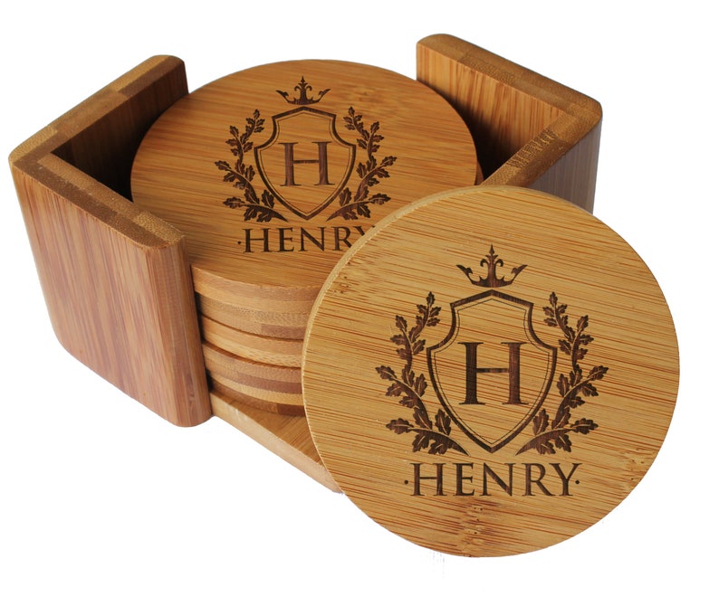 May include: A set of round light brown bamboo coasters stored in a matching square wooden holder. Each coaster features a laser-engraved design of a shield with the letter H inside, topped with a crown and flanked by oak leaves, with the name HENRY below.