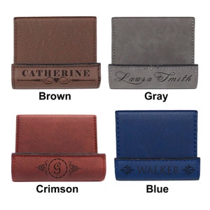 May include: Four leather business card holders in different colors: brown, gray, crimson, and blue. Each holder has a different engraved design. The brown holder has the name "Catherine" engraved on it. The gray holder has the name "Laura Smith" engraved on it. The crimson holder has the letter "G" engraved on it. The blue holder has the word "WALKER" engraved on it.