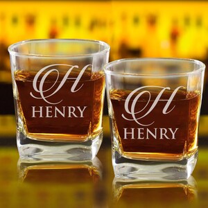 May include: Two clear glass whiskey tumblers with a square base. Each glass is engraved with a cursive "H" and the name "HENRY". The glasses are filled with amber liquid.