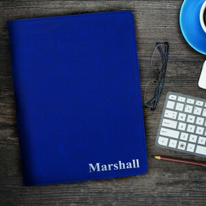 May include: A royal blue leather-like portfolio folder featuring the name Marshall embossed in silver lettering on the bottom right corner. The rectangular folder has a smooth, solid-colored surface and is closed, showcasing a clean, professional design.