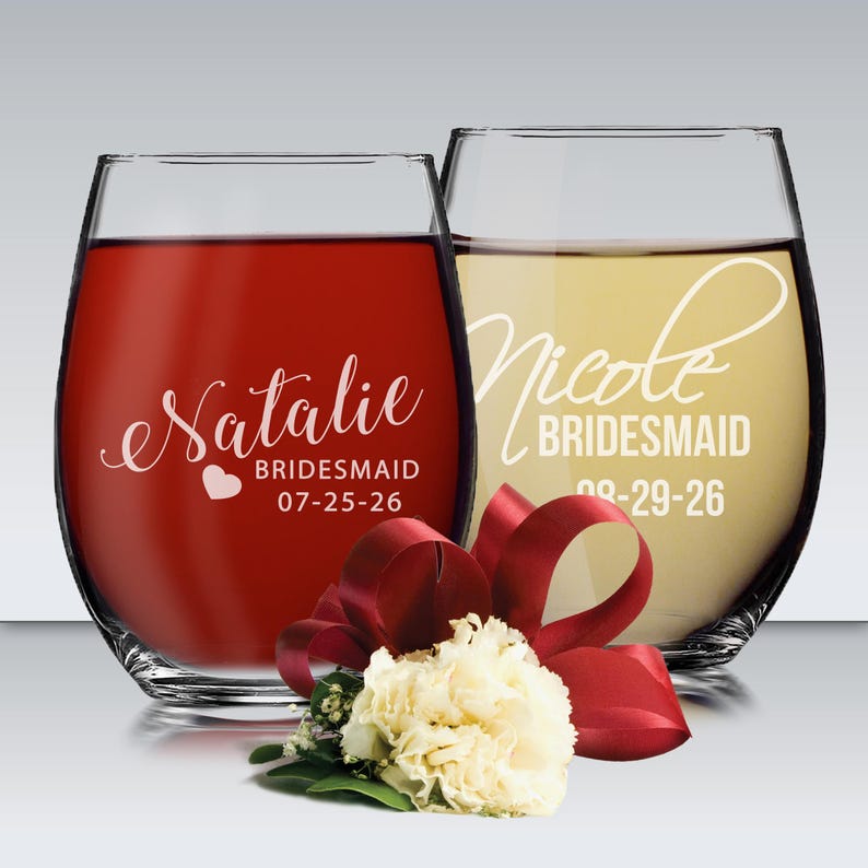 May include: Two stemless wine glasses, one filled with red liquid and the other with a yellow liquid. The red glass has the name "Natalie", the word "Bridesmaid", and a date. The yellow glass has the name "Nicole", the word "Bridesmaid", and a date. A red ribbon and white flower are in front.