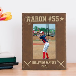 May include: Brown leather photo frame with a baseball theme. The frame has a photo of a young baseball player in a red, white, and blue uniform. The frame is engraved with the text "AARON #55", "BELLEVUE RAPTOR", and "2023".