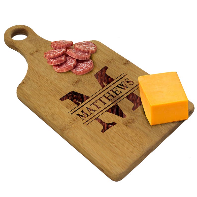 May include: A personalized bamboo cutting board with a handle. The board has the letter "M" engraved on it, along with the name "MATTHEWS". There are slices of salami and a block of cheddar cheese on the board.