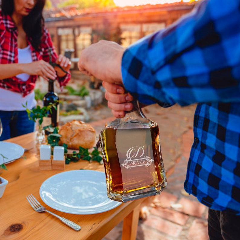 May include: A clear glass decanter filled with amber liquid, featuring an etched design with the letter D and the name DRAKE. The square-shaped vessel has a thick base and is held by a person wearing a blue and black plaid flannel shirt.