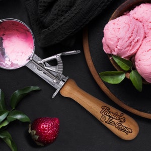 May include: A wooden ice cream scoop with a silver handle and the text "Tamia's Ice Cream" engraved on the handle. The scoop is next to a bowl of pink ice cream and a strawberry.
