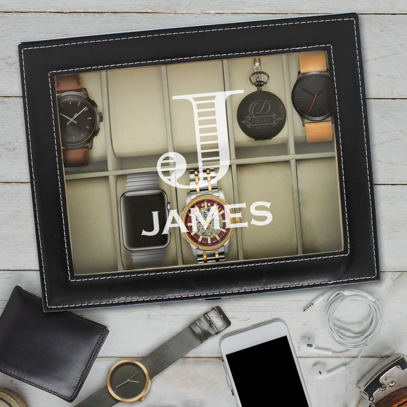 May include: A black rectangular watch display case with white stitching and a glass lid featuring the white letter J and the name JAMES. The interior has beige compartments holding various wristwatches and a pocket watch with a tan strap.