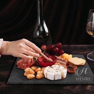 May include: A rectangular black slate serving board engraved with the letter H and the name HENRY. The board holds a spread of brie cheese, crackers, salami, dried figs, grapes, and a small red bowl containing honey with a wooden honey dipper.