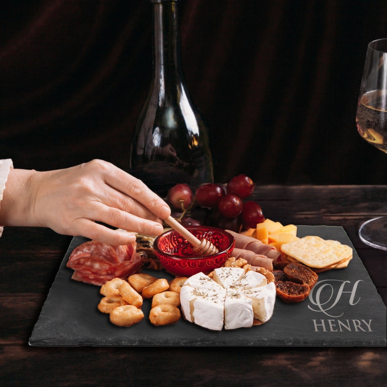 May include: A rectangular black slate serving board engraved with the letter H and the name HENRY. The board holds a spread of brie cheese, crackers, salami, dried figs, grapes, and a small red bowl containing honey with a wooden honey dipper.