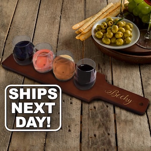May include: A dark brown wooden serving paddle with the name Becky engraved in script, holding four stemless glass tumblers filled with various colored wines, accompanied by a small white bowl of green olives and breadsticks.