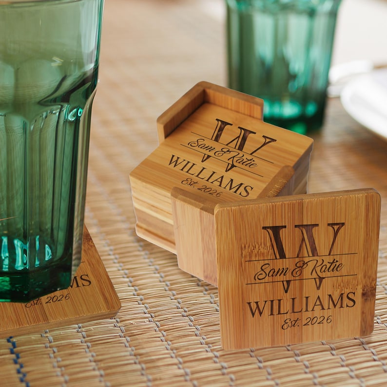 May include: A set of square bamboo coasters with a holder, featuring a laser-engraved design with the letter W, the names Sam & Katie, the surname WILLIAMS, and the text Est. 2026. The coasters have a natural wood grain finish and light brown color.