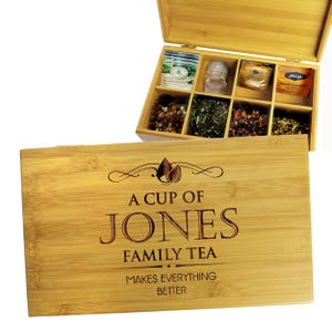 May include: A bamboo tea box with a hinged lid, containing various tea bags and loose-leaf teas. The box is engraved with the text "A CUP OF JONES FAMILY TEA MAKES EVERYTHING BETTER". The box is made of light brown bamboo.