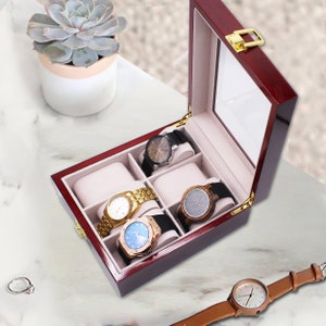 Pode incluir: Uma caixa de rel&oacute;gio de madeira vermelha escura brilhante, com cinco rel&oacute;gios com diferentes pulseiras e mostradores. A caixa tem uma tampa de vidro e um fecho dourado. Uma planta suculenta num vaso branco est&aacute; no fundo.