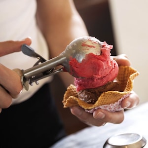 May include: A person scoops pink and brown ice cream into a waffle cone with a silver ice cream scoop.
