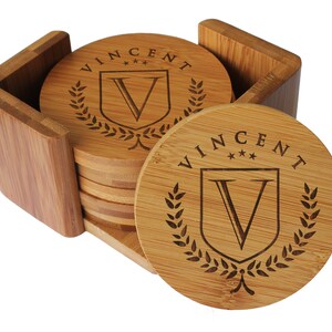 May include: A set of round bamboo coasters stacked inside a matching square bamboo holder. Each coaster features a laser-engraved design with the name VINCENT, three small stars, a shield containing the letter V, and a laurel wreath border.