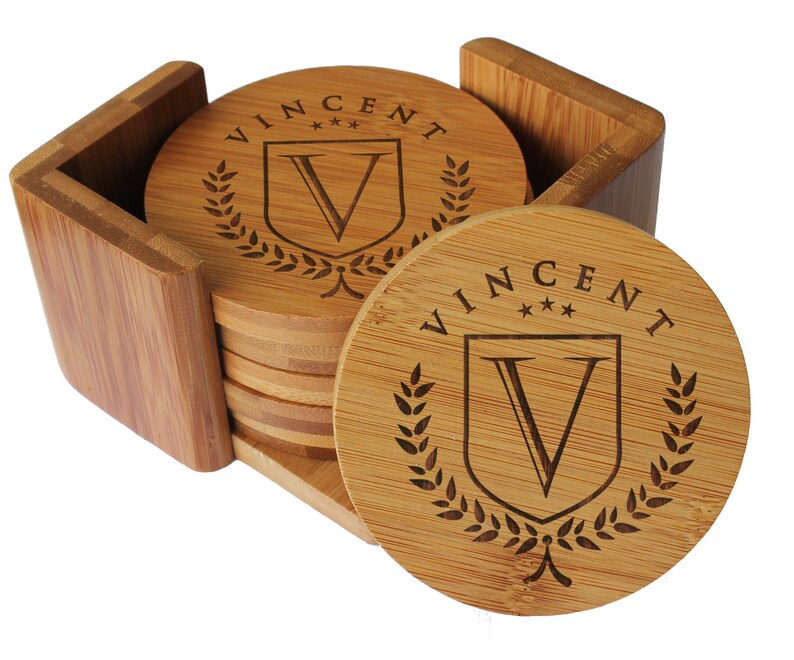 May include: A set of round bamboo coasters stacked inside a matching square bamboo holder. Each coaster features a laser-engraved design with the name VINCENT, three small stars, a shield containing the letter V, and a laurel wreath border.