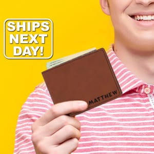 May include: A brown leather bifold wallet featuring the name MATTHEW engraved in black lettering on the bottom right corner. The wallet is shown holding paper currency inside its main compartment, with visible dark stitching along the edges.