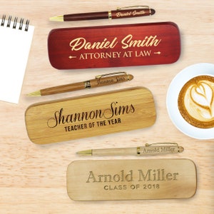 May include: Three wooden boxes with engraved names and titles, each containing a pen. The boxes are on a wooden surface. The names are Daniel Smith, Attorney at Law, Shannon Sims, Teacher of the Year, and Arnold Miller, Class of 2018.